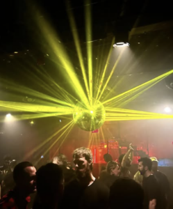 A lively crowd dances in a dimly lit LGBTQ nightclub, with yellow beams of light radiating from a central disco ball above.